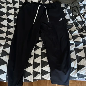 Svarta byxor från Nike - Säljer ett par svarta mjukisbyxor från Nike med vit snörning i midjan. Byxorna har en avslappnad passform och är perfekta för en bekväm stil. Nike-loggan är tryckt på ena benet.