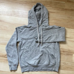 Grå hoodie från H&M - Säljer en klassisk grå hoodie från H&M Basics. Den har en stor ficka framtill och justerbar huva med snören. Perfekt för en avslappnad stil.