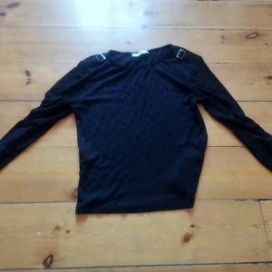 Svart långärmad topp från Mango - Säljer en stilren svart långärmad topp från Mango. Toppen har dekorativa spännen på axlarna och är tillverkad i ett ribbat material som ger en snygg textur. Perfekt för en chic och enkel look.