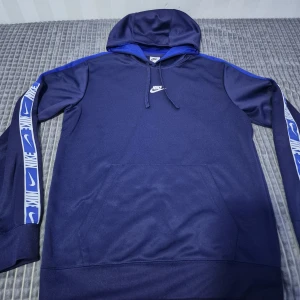 Nike mörkblå hoodie  - Säljer en snygg mörkblå hoodie från Nike med vita logotyper längs ärmarna och på bröstet. Hoodien har en justerbar huva och en stor ficka framtill. Perfekt för en sportig look!