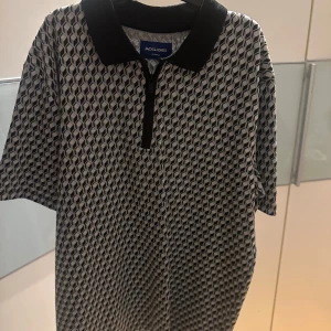 Mönstrad pikétröja från Jack & Jones - Snygg pikétröja från Jack & Jones med ett geometriskt mönster i svart och grått. Tröjan har en klassisk krage och korta ärmar.