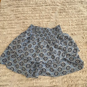 Blå mönstrad kjol från Gina Tricot - Säljer en söt blå kjol från Gina Tricot med ett fint mönster i vitt och mörkblått. Kjolen har en elastisk midja och volangdetaljer som ger en lekfull look. Perfekt för varma dagar!