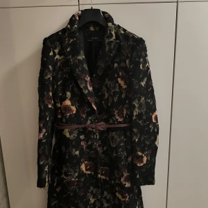 Blommig kappa från Zara Basic - Snygg kappa från Zara Basic med ett blommigt mönster i dova färger. Den har ett bälte i midjan och långa ärmar. Perfekt för att ge din outfit en elegant touch.