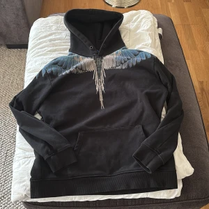 Svart hoodie från Marcelo Burlon - Säljer en svart hoodie från Marcelo Burlon med ett coolt vingmönster i blått och vitt över axlarna. Tröjan har en klassisk passform med långa ärmar och en stor ficka framtill. Perfekt för en stilren och avslappnad look.