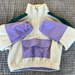 Beige fleecejacka med lila detaljer - Säljer en mysig beige fleecejacka med lila och gröna detaljer. Jackan har en halv dragkedja och en stor ficka framtill. Perfekt för kyliga dagar!