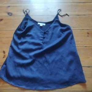 Svart linnetopp från Amisu - Snygg svart linnetopp från Amisu med smala axelband och v-ringning. Perfekt för varma dagar eller som ett lager under en kavaj. Tillverkad i ett mjukt och lätt material som ger en skön känsla mot huden.