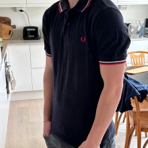 Svart pikétröja från Fred Perry - Säljer en klassisk svart pikétröja från Fred Perry med röd och vit rand på krage och ärmslut. Tröjan har en broderad logga på bröstet och korta ärmar. Perfekt för en stilren look!