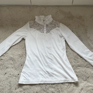 Vit långärmad topp med spetsdetaljer - Elegant vit långärmad topp med spetsdetaljer på framsida och rygg. Har en hög krage med volang och knappar framtill. Perfekt för tävling. Nypris: 499kr Mitt pris 149kr + frakt. Kontakta gärna innan köp nu, säljer en likadan utan gul text❣️