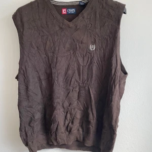 Brun väst från Chaps - Säljer en brun väst från Chaps med v-ringad design. Västen har ribbade kanter och ett broderat emblem på bröstet. Perfekt för lager-på-lager under kyligare dagar.