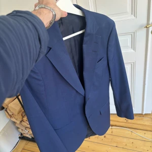 Blå kavaj från Tiger of Sweden - Snygg blå kavaj från Tiger of Sweden med klassisk design. Kavajen har en enkelknäppning och fyra knappar vid ärmsluten. Använd vid ett tillfälle, därav nyskick! 