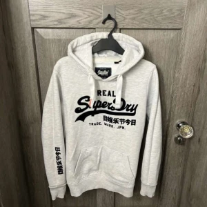 Grå hoodie från Superdry - Säljer en stilren grå hoodie från Superdry med svart tryck på bröstet och ärmen.  Storlek s 