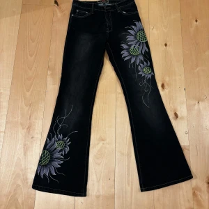 Svarta jeans med blombroderi - Unika svarta jeans med vackert broderade blommor i lila och grönt längs benen. Byxorna har en bootcut-stil som ger en snygg siluett. Perfekta för att ge din outfit en kreativ touch. Strl står inte på byxorna men skulle gissa runt xs-s. Aldrig använda🩷