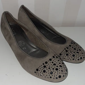 Grå ballerinaskor med dekorativa stenar nya  - Snygga gråa ballerinaskor från Ara med dekorativa svarta stenar på tån. Skorna är tillverkade i mocka och har en låg klack för extra komfort. Perfekta för en stilren look.