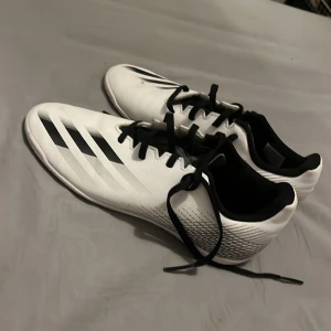 Vita fotbollsskor från Adidas - Snygga vita fotbollsskor från Adidas med svarta detaljer och snörning. Perfekta för inomhusspel med en slitstark sula. Skorna har en modern design med klassiska ränder på sidan.