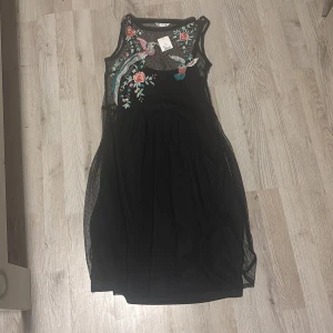Svart klänning med broderade blommor och fåglar - Säljer en svart klänning som inte har använt med vackra broderade blommor och fåglar i rosa, blått och grönt. Klänningen är ärmlös och har ett transparent mesh-överdrag som ger en elegant look. Perfekt för speciella tillfällen!