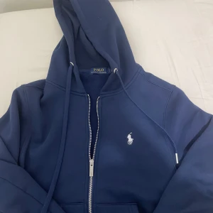 Mörkblå hoodie från Ralph Lauren - Säljer en snygg mörkblå hoodie från Ralph Lauren med dragkedja och det klassiska polospelar-emblemet på bröstet. Perfekt för en stilren och avslappnad look. Huvan har justerbara snören för bästa passform.