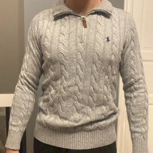 Grå stickad tröja från Ralph Lauren - Säljer en stilren grå stickad tröja från Ralph Lauren med kabelstickat mönster och dragkedja vid halsen. Tröjan har långa ärmar och en broderad logotyp på bröstet. Perfekt för kyliga dagar!