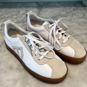 Vita sneakers från Blankens strl 37 - Snygga vita sneakers med beige detaljer från Blankens. Skorna har snörning och en stilren design som passar perfekt för en avslappnad look. Tillverkade i en kombination av skinn och mocka för en lyxig känsla. Helt nya och oanvända. Säljes pga fel storlek. Nypris 2800