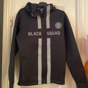 Svart hoodie från Black Squad - Säljer en svart hoodie från Black Squad med texten 'BLACK SQUAD' och ett tryck av en jordglob på bröstet. Hoodien har en dragkedja framtill och en justerbar huva. Perfekt för en casual look.