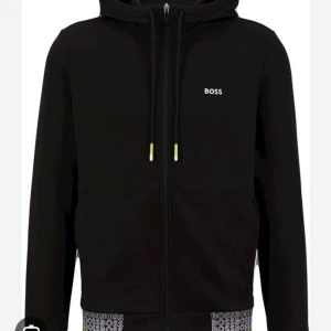 Svart hoodie från Boss - Säljer en stilren svart hoodie från Boss med dragkedja och justerbar huva. Tröjan har en diskret logotyp på bröstet och ribbade muddar vid ärmslut och nederkant. Perfekt för en avslappnad stil.