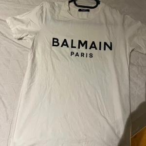 Balmain Paris T Shirt - Säljer en stilren vit t-shirt från Balmain med svart text 'BALMAIN PARIS' på framsidan. T-shirten är kortärmad och tillverkad i Portugal. Perfekt för en casual look. Nypris ca 2500 kr 