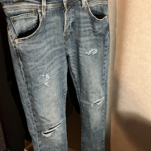 Blå jeans från Jack & Jones - Snygga blå jeans från Jack & Jones med slitna detaljer och en slim passform. De har en klassisk femficksdesign och är perfekta för en avslappnad stil. Märket 'Slim Glenn' syns på insidan av midjan.