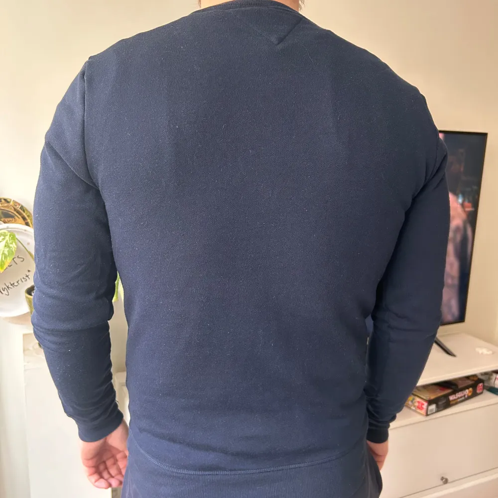 Säljer en stilren mörkblå tröja från Tommy Hilfiger med märkets logga på bröstet och text längs ärmarna. Tröjan har långa ärmar och en rund halsringning. Perfekt för en casual look. Modellen är 185 cm och väger 100 kg. . Neuletakit & Villapaidat.