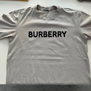 Grå t-shirt från Burberry - Säljer en stilren grå t-shirt från Burberry med deras ikoniska logga tryckt i svart på bröstet. Perfekt för en casual look. T-shirten har en normal passform och är kortärmad. Passar utmärkt till både jeans och shorts.