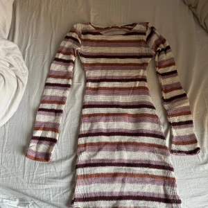 Randig stickad strandklänning - Säljer en randig stickad strandklänning i härliga toner av lila, beige och brun. Klänningen har en öppen rygg och långa ärmar, perfekt för en avslappnad stil. Den är gjord i ett mjukt och bekvämt material. Klänningen är i S men är mer som en XS