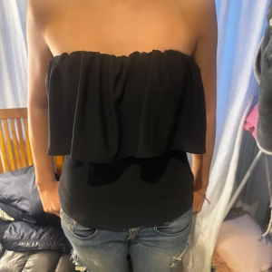 Svart axelbandslös topp - Säljer en stilren svart axelbandslös topp med volangdetalj. Perfekt för en kväll ute eller en somrig dag. Toppen har en tight passform och är tillverkad i ett mjukt material som sitter bekvämt.