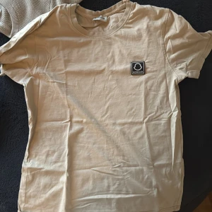 Beige t-shirt från Moncler - Säljer en stilren beige t-shirt från Moncler med en liten logotyp på bröstet. T-shirten har en normal passform och är kortärmad, perfekt för en avslappnad look. Tillverkad i mjukt material för extra komfort.