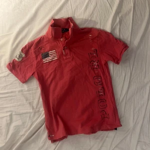 Röd pikétröja från Ralph Lauren - Säljer en röd pikétröja från Ralph Lauren med korta ärmar och tryck på både fram- och baksidan. Tröjan har en klassisk krage och knappar framtill. Perfekt för en avslappnad stil. Skriv vid minsta funderingar!👍