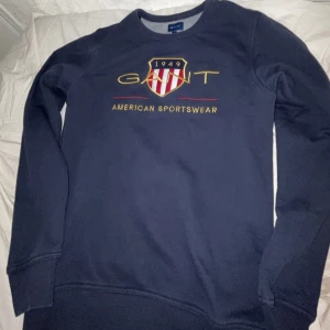 GANT - Säljer en stilren mörkblå sweatshirt från GANT med tryck framtill. Tröjan har långa ärmar och rund halsringning. Perfekt för en avslappnad stil.