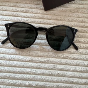 Svarta solglasögon från Oliver Peoples - Snygga svarta solglasögon från Oliver Peoples med en klassisk rund form. De har mörka glas och en stilren design. Perfekta för soliga dagar och ger en tidlös look.