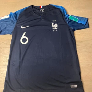 Blå fotbollströja från Nike - Säljer en blå fotbollströja från Nike med korta ärmar. Tröjan har ett tryck med nummer 6 och texten 'POGBA' på ryggen. Den har även ett emblem på bröstet och en Nike-logga. Perfekt för fotbollsfans!