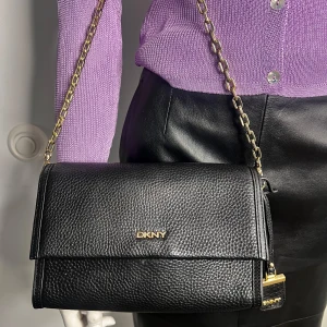 Svart axelväska från DKNY - Snygg svart axelväska från DKNY i skinn med guldfärgade kedjedetaljer. Väskan har en elegant design med logotyp framtill och en praktisk axelrem. Perfekt för att ge en stilren touch till din outfit. #DKNY #väska 