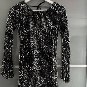 Svart glittrig klänning - Snygg svart klänning täckt med glittriga paljetter. Den har långa ärmar och en djup öppen rygg med knytband. Perfekt för en festlig kväll! Den är kort.