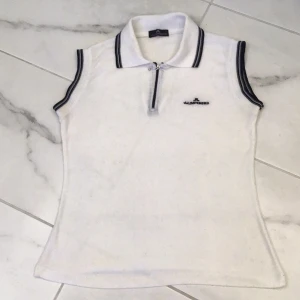 Vit ärmlös pikétröja från J.Lindeberg - Snygg vit ärmlös pikétröja från J.Lindeberg med dragkedja och broderad logga på bröstet. Tröjan har svarta detaljer på krage och ärmöppningar. Perfekt för en stilren look!