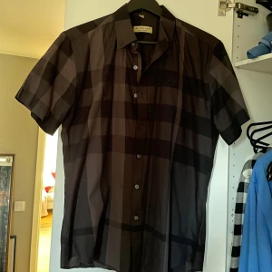 Svart och grå rutig skjorta från Burberry - Snygg kortärmad skjorta från Burberry i svart och grå med klassiskt rutmönster. Skjortan har knappar framtill och en diskret broderad logga på bröstet. Perfekt för en stilren look.