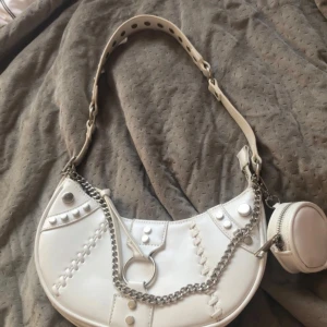 Vit axelväska med kedjedetalj - Snygg vit axelväska i syntetmaterial med coola kedjedetaljer och nitar. Väskan har en unik form och en liten avtagbar väska med dragkedja. Perfekt för att ge din outfit en edgy touch!
