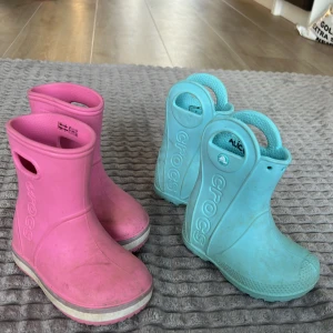 Rosa och turkosa stövlar från Crocs - Säljer ett par färgglada Crocs-stövlar i rosa och turkos. Perfekta för regniga dagar med deras vattentäta design och bekväma passform. Lätta att ta på och av med praktiska handtag.