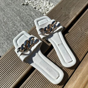 Vita sandaler med kedjedetalj från Zara - Säljer ett par trendiga vita sandaler från Zara med fyrkantig tå och bred rem framtill. Remmen är dekorerad med en stor guldfärgad kedja som ger en cool touch. Platt sula och öppen tå, perfekta för sommaren.