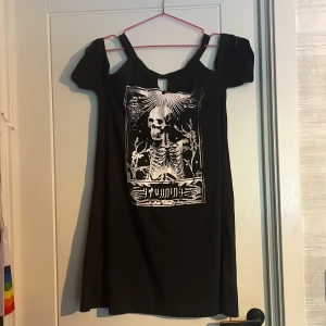 Svart kortklänning med skelett-tryck från SHEIN - Svart kortklänning från SHEIN med vitt skelett-tryck framtill. Klänningen har cold shoulder-design med smala axelband och korta ärmar. Perfekt för dig som gillar alternativ stil och vill sticka ut.
