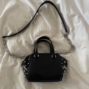 Svart handväska med nitar och kedjedetaljer - Cool svart handväska med silverfärgade nitar på sidorna och en grov kedja som axelrem. Väskan har både handtag och axelrem samt dragkedja upptill. 