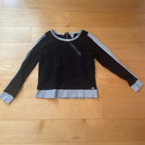 Svart och grå sweatshirt från Guess - Säljer en svart sweatshirt från Guess med gråa muddar och detaljer vid ärmslut, nederkant och axlar. Tröjan har långa ärmar och en klassisk rund halsringning. Perfekt för en avslappnad och stilren look. Köparen står för frakten!