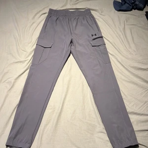 Grå cargobyxor från Under Armour - Säljer ett par grå cargobyxor från Under Armour med elastisk midja och muddar vid benslut. Byxorna har två stora fickor med lock på sidorna och är perfekta för en sportig och avslappnad stil.