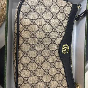 Beige och svart axelväska från Gucci - Säljer en beige axelväska från Gucci med klassiskt GG-mönster och svarta skinn-detaljer. Väskan har guldfärgad logga och dragkedja upptill. Perfekt för dig som vill ha en stilren och lyxig accessoar.