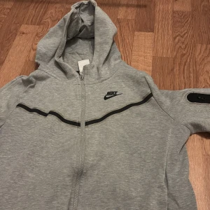 Grå zip-hoodie från Nike - Säljer en grå hoodie från Nike med dragkedja och huva. Jackan har svarta detaljer och en svart Nike-logga på bröstet. Perfekt för en sportig och avslappnad stil.