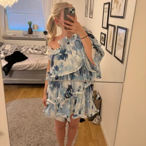 Klänning från lojsan wallins kollektion  - Säljer en superfin kort klänning i blått och vitt med blommigt mönster från lojsan Wallins kollektion med nakd. Klänningen har volangdetaljer, omlott-design och är oneshoulder. Klänningen är använd fåtal gånger och säljer då den inte kommer till användning 