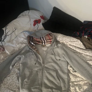 Grå hoodie med dragkedja från Burberry - Säljer en grå hoodie från Burberry med dragkedja och klassiskt rutigt foder i huvan. Jackan har två fickor framtill och justerbar huva med snörning. Perfekt för dig som gillar stilrena plagg med en twist.